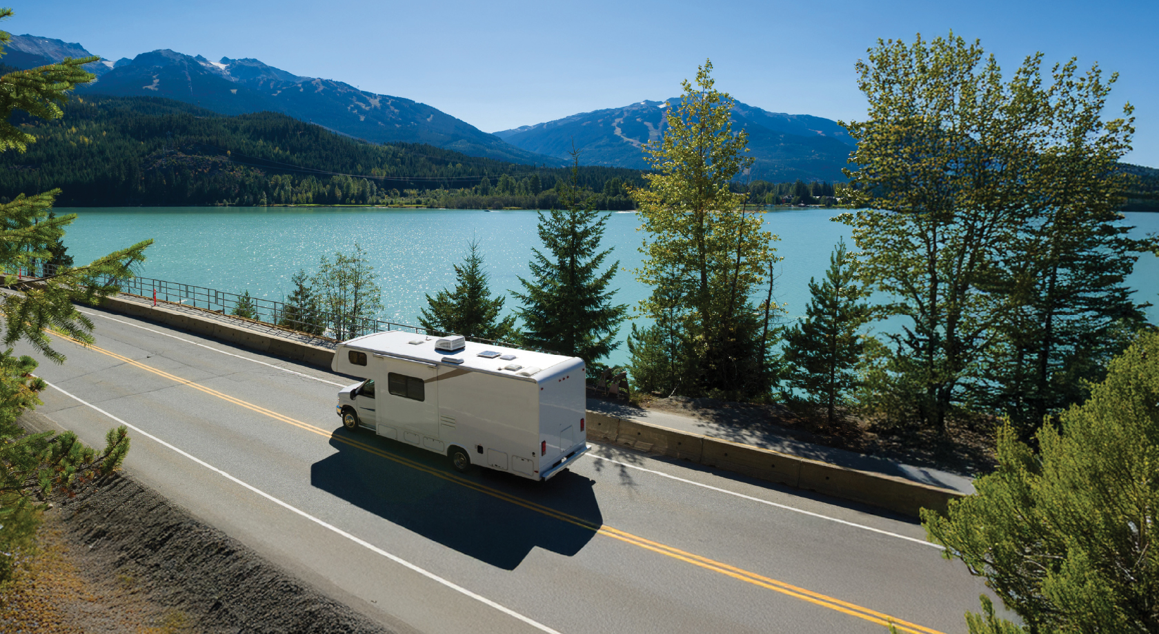 Image of White RV driving on highway next to mountain and lake scenery. 